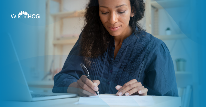 Image of Black woman writing notes in a home office with a laptop next to her