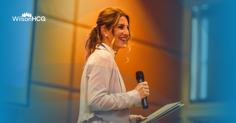 A lady on stage holding a microphone wearing a beige jumper